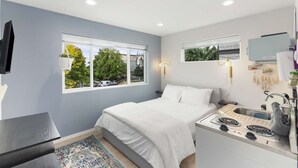 A compact bedroom with a bed, a small desk, a built-in stove, and two windows with blinds. - Anders' sweet suite - Micro Studio - 91Walkscore (Seattle)