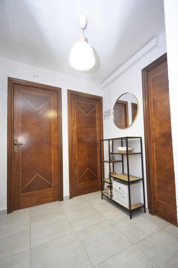 Un couloir avec deux portes en bois, un luminaire blanc au plafond, un miroir et une étagère en métal noir sur laquelle se trouve une boîte blanche.
