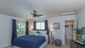 Une chambre à coucher comprenant un lit, un ventilateur de plafond, une fenêtre avec des rideaux, une table de chevet avec une lampe et un panier.