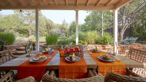 Un patio al aire libre con zona de comedor cubierta, con mesas dispuestas con platos, vasos y servilletas, rodeado de sillas de mimbre y un muro de piedra.