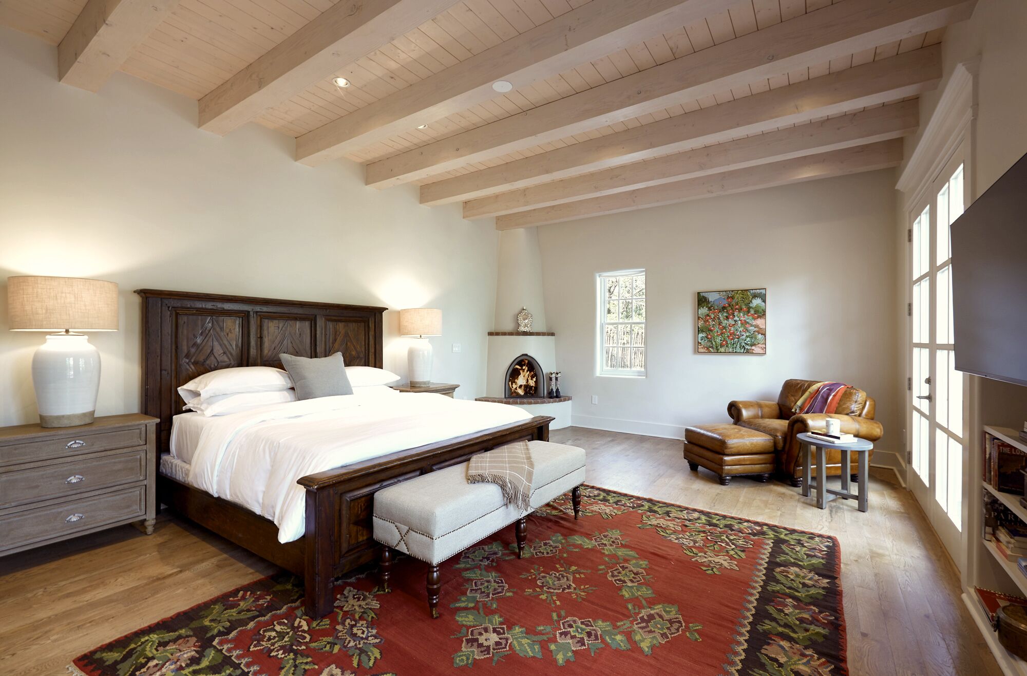 A bedroom with a large bed, bedside lamps, a bench, a fireplace, a chair, a rug, and a bookshelf.