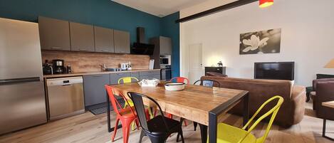 A modern kitchen with stainless steel appliances, a wooden dining table with colorful chairs, and a living area with a sofa and television.