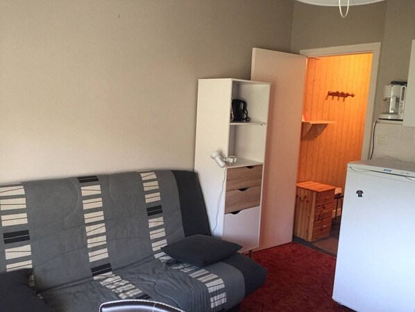 A compact room with a grey sofa, a white wardrobe, a small wooden cabinet, and a white refrigerator.