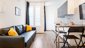 A modern apartment with a black leather sofa, a dining table with chairs, a kitchen area, and a large window with curtains.