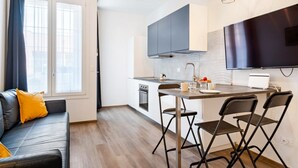 A modern kitchen with a dining area, a sofa with cushions, and a TV mounted on the wall.