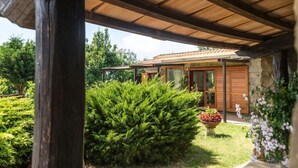 Une structure en bois dotée d’un patio couvert, un bâtiment en pierre avec des portes vitrées et un jardin bien entretenu agrémenté d’arbustes et d’un pot de fleurs.