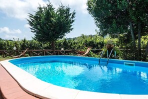 Une piscine rectangulaire dotée d’une terrasse en bois et d’un escalier en métal, entourée d’une végétation luxuriante et d’arbres.