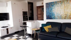 Un espace de vie compact comprenant un coin cuisine, un canapé noir agrémenté de coussins à motifs, un petit tabouret de bar et un grand tableau abstrait accroché au mur.