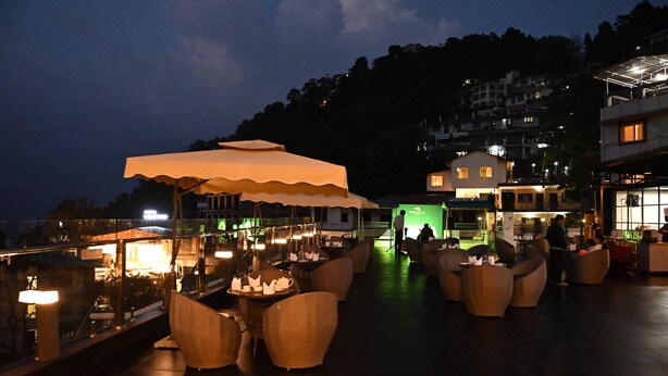 An outdoor seating area with wicker furniture and umbrellas, a staircase leading to a building, and a view of a hillside. - Pride Ashiyana Resort Mussoorie (Dehradun)