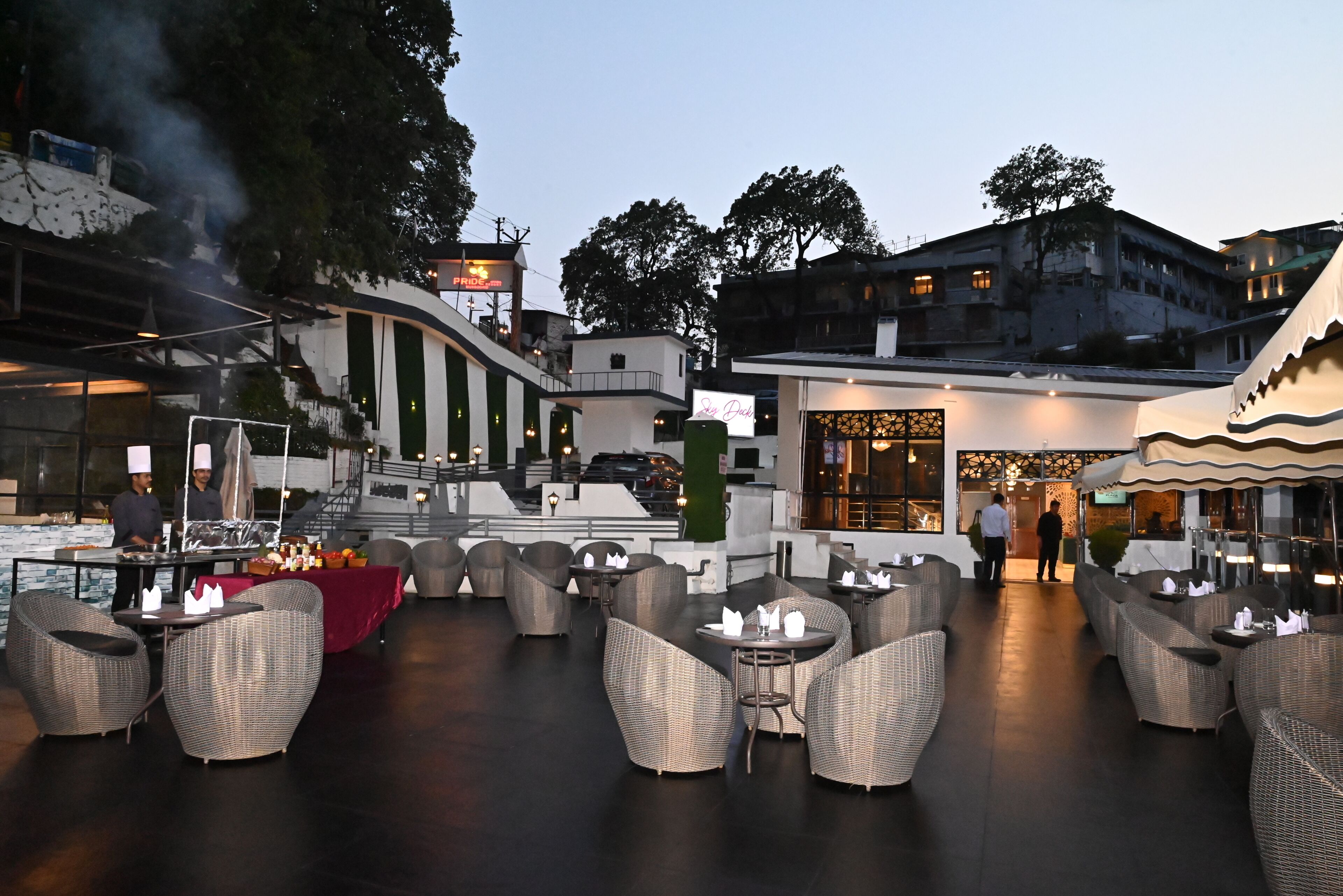 An outdoor dining area with wicker furniture and a red tablecloth, surrounded by buildings and trees.