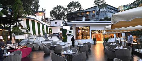 An outdoor dining area with wicker chairs and tables, a buffet setup, and a building with a large entrance.