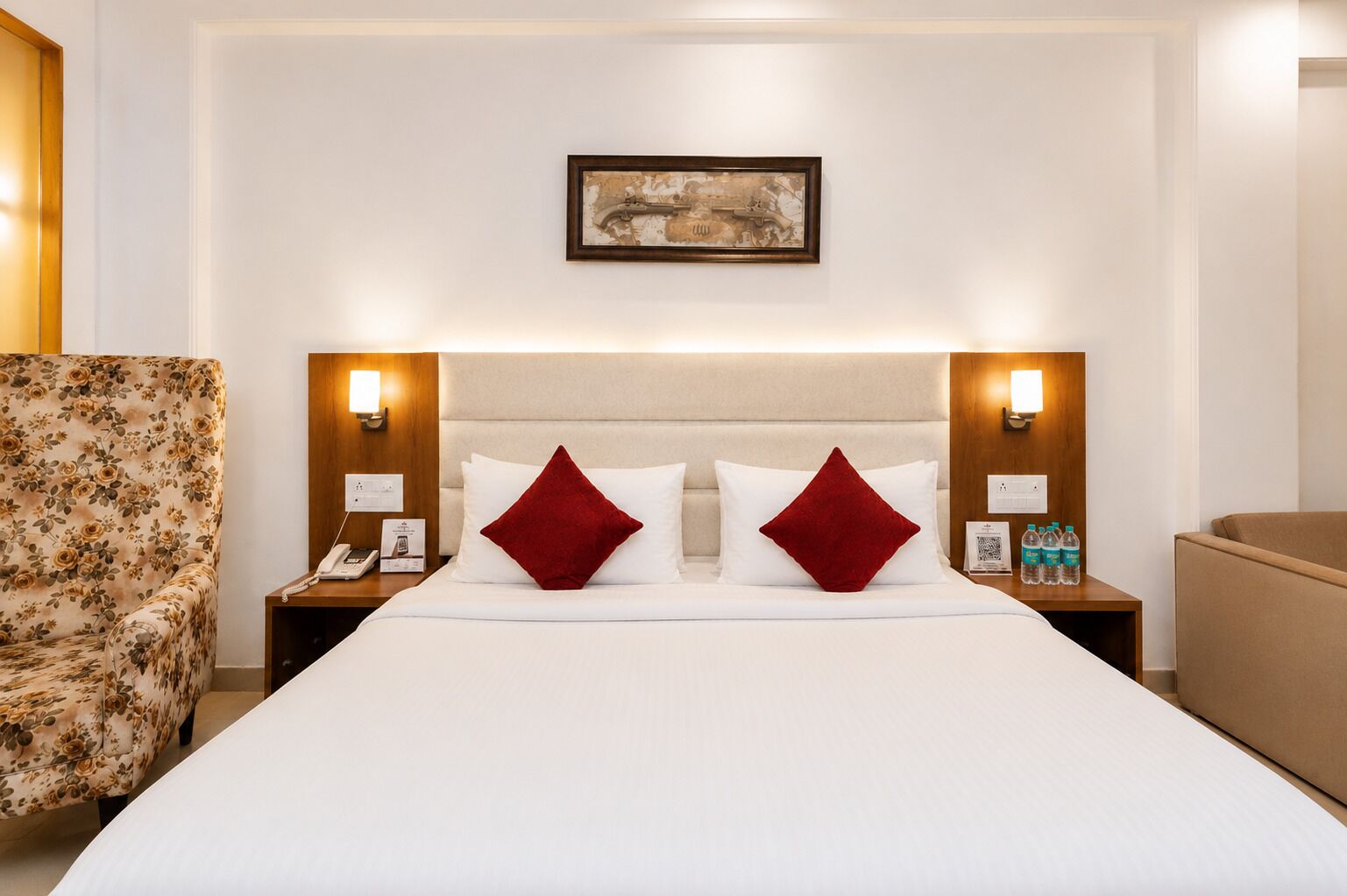 A hotel room with a large bed, two nightstands, a floral armchair, and a framed picture on the wall.
