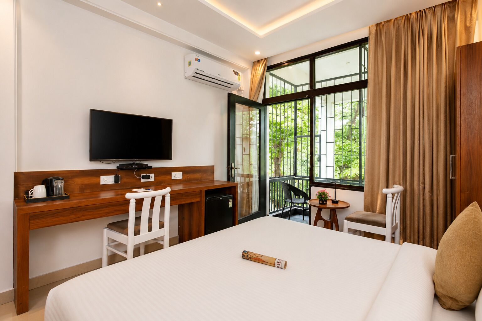 A hotel room with a bed, a desk with a television, a small table with a plant, and a balcony with curtains.