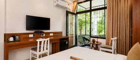 A hotel room with a bed, a desk with a television, a small table with a plant, and a balcony with curtains.