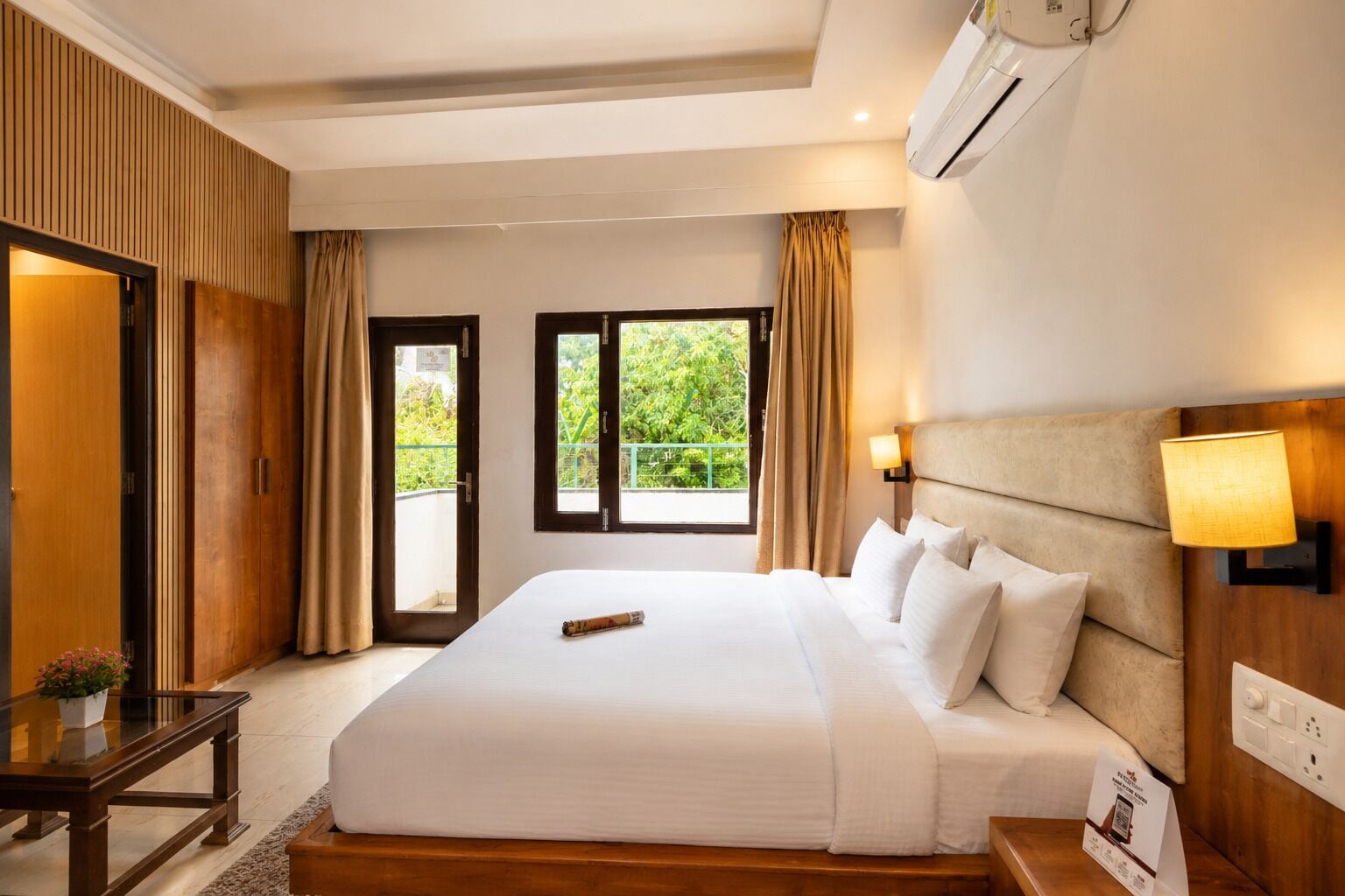 Quarto de hotel com uma cama grande, escrivaninha, cadeira, abajur e vista para o verde pela janela.