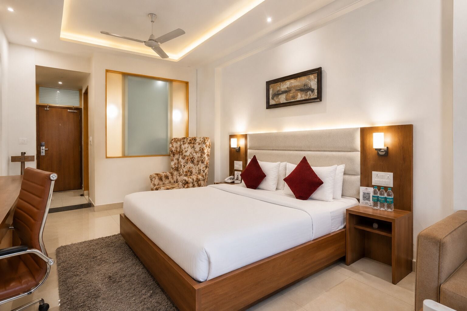 A hotel room with a large bed, a desk, a chair, a ceiling fan, and a painting on the wall.