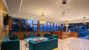 A spacious lounge with teal sofas, a central glass coffee table, and large windows offering a view of the mountains. - Pride Ashiyana Resort Mussoorie (Dehradun)