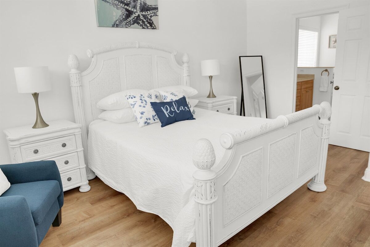 A white four-poster bed with a textured headboard, two bedside tables with lamps, a blue accent pillow, and a blue chair.