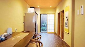 A narrow hallway with a wooden table and chairs, a refrigerator, and a sliding glass door leading to a balcony with a view of trees. - INN Plus Toji Kindo (Kyoto)