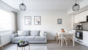 Un salon moderne avec un canapé gris, une table à manger avec des chaises et un coin cuisine doté de meubles blancs et d’une cuisinière.