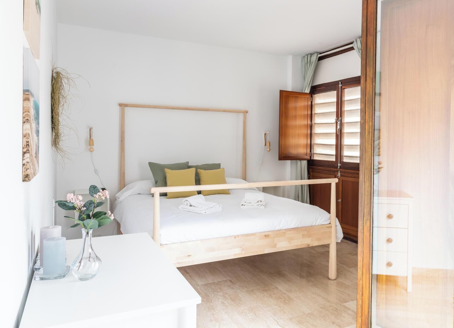 A neatly arranged bedroom with a wooden bed frame, a bedside table with a vase and candles, and a wardrobe with a window.