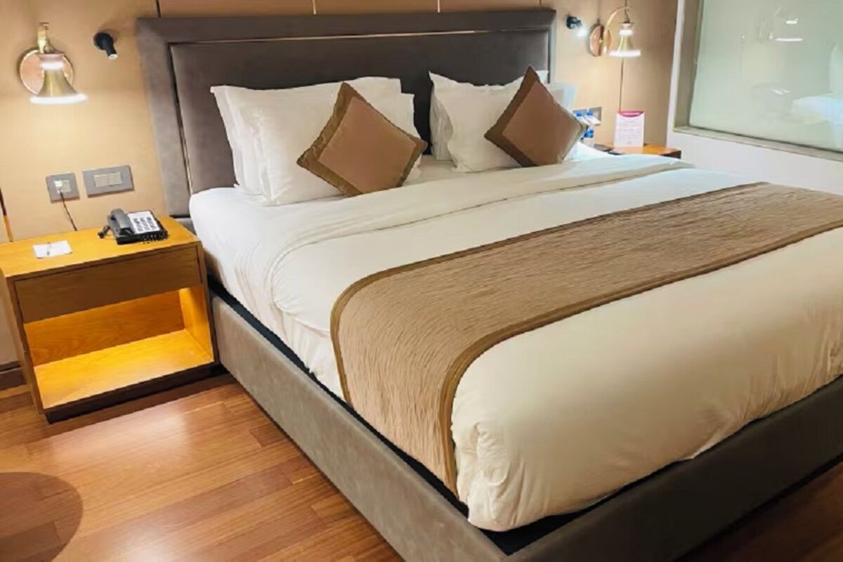 Quarto de hotel com uma cama grande, uma mesa de cabeceira de madeira, abajur e vista da cidade pela janela.