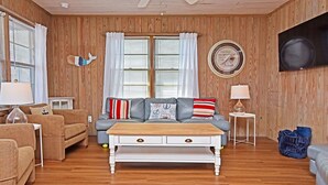 Ein Wohnzimmer mit Holzbalkendecke, einem Deckenventilator, einem grauen Sofa mit gestreiften Kissen, einem weißen Couchtisch und einem an der Wand montierten Fernseher.