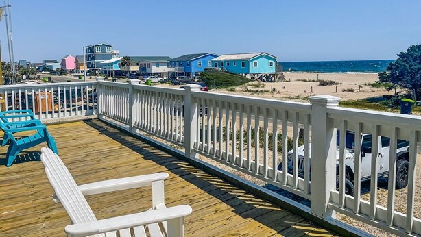 Eine Holzterrasse mit weißer Reling, die auf einen Strand mit Häusern und einem strahlend blauen Himmel blickt.