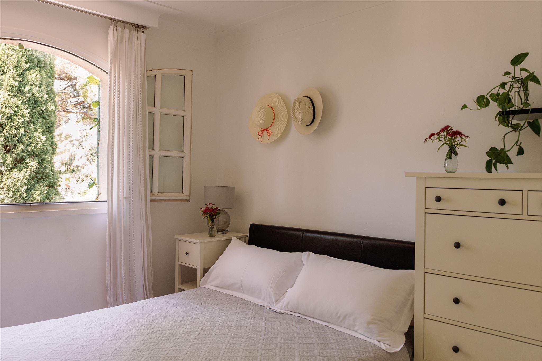 Un dormitorio con cama, mesita de noche, cómoda y dos sombreros colgados en la pared.