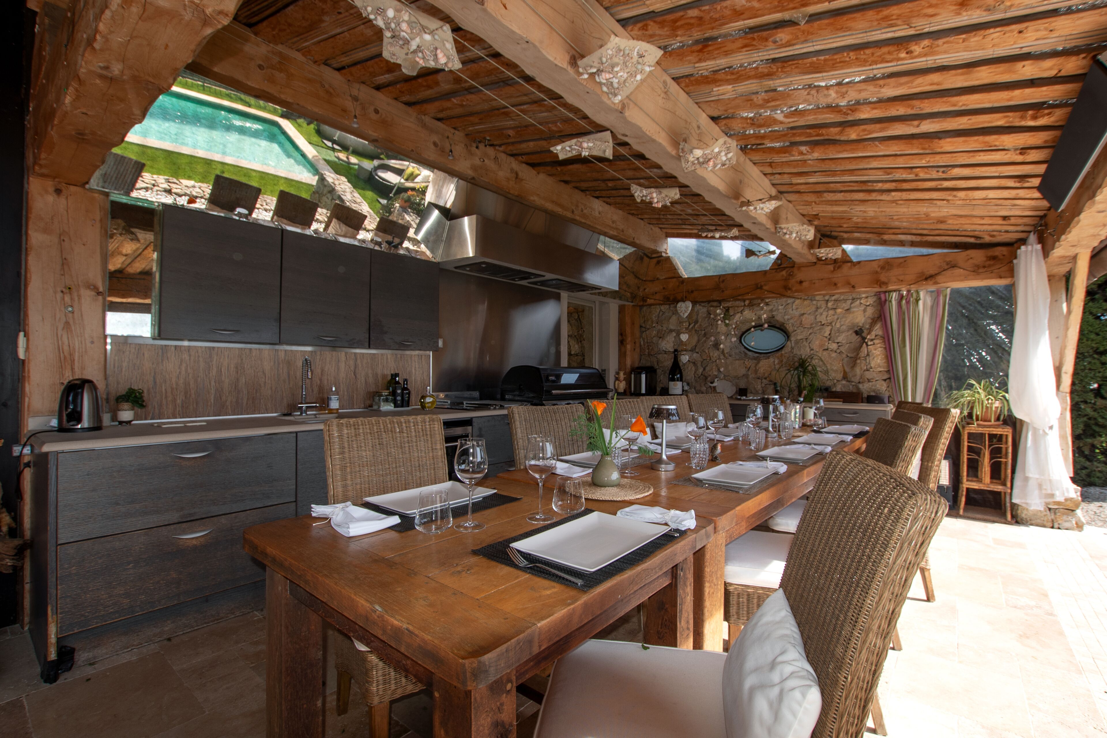 A wooden dining area with a long table set for four, surrounded by wicker chairs. A kitchen with modern appliances and a stone wall is visible in the background.