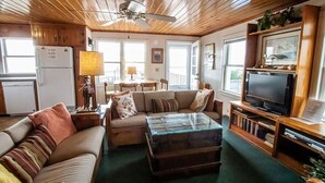 A cozy living room with a wooden ceiling, a glass coffee table, a sofa, and a television on a wooden stand. - Experience oceanfront bliss at The Captain's Quarters, Holden Beach! (Holden Beach)