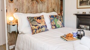 A bedroom with a wooden headboard, floral pillows, a bedside table with a lamp, a fireplace, and a painting on the wall. - Michaelmass Cottage (Whitby)