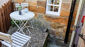 A stone-walled outdoor area with a round table and chairs, a small window, and a wooden fence. - Michaelmass Cottage (Whitby)