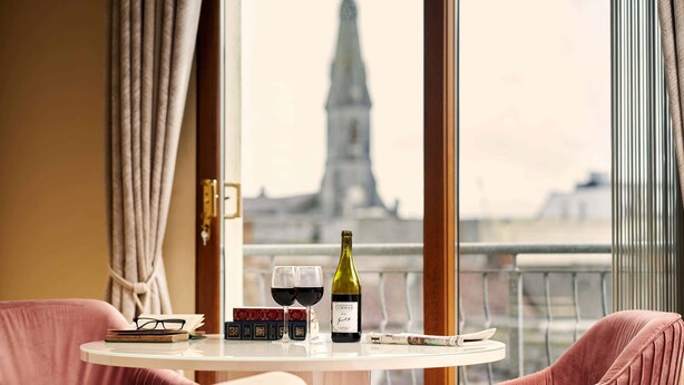 Una mesa con una botella de vino, dos copas y libros, con vistas a una torre a través de amplios ventanales.
