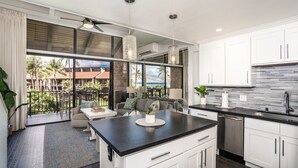 A modern kitchen with white cabinets, a black countertop, and a large island. There is a dining area with a sofa and a table, and a balcony with a view of palm trees and a building.