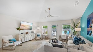 Un salón amplio con un ventilador de techo blanco, un televisor de pantalla plana grande y vistas al exterior a través de las ventanas.