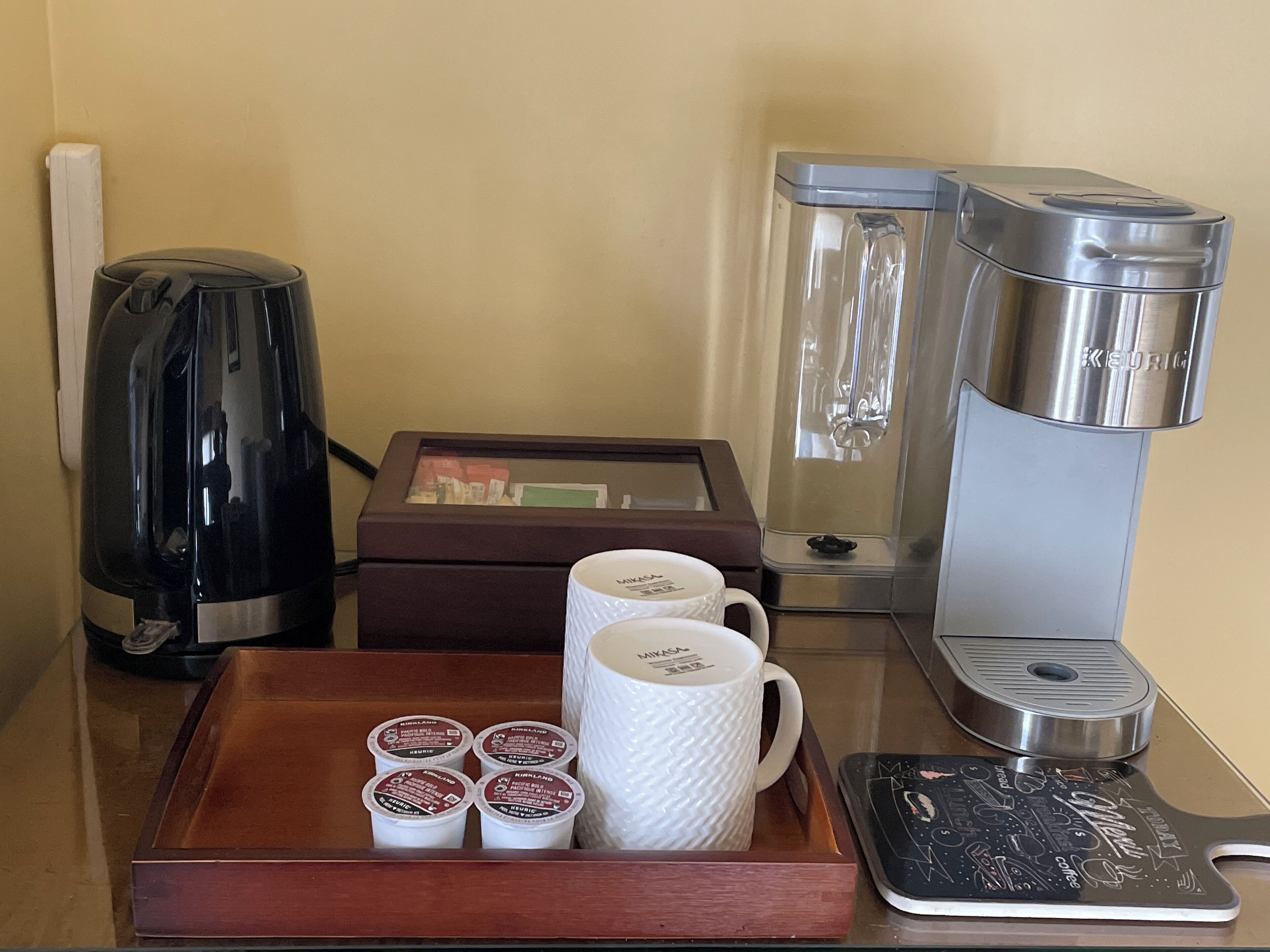 Estação de café com cafeteira, chaleira e bandeja com xícaras de café e embalagem de cápsulas.