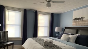 A bedroom with a large bed, two windows with curtains, a ceiling fan, a nightstand with a lamp, and a painting on the wall. - The York House (Fort Erie)