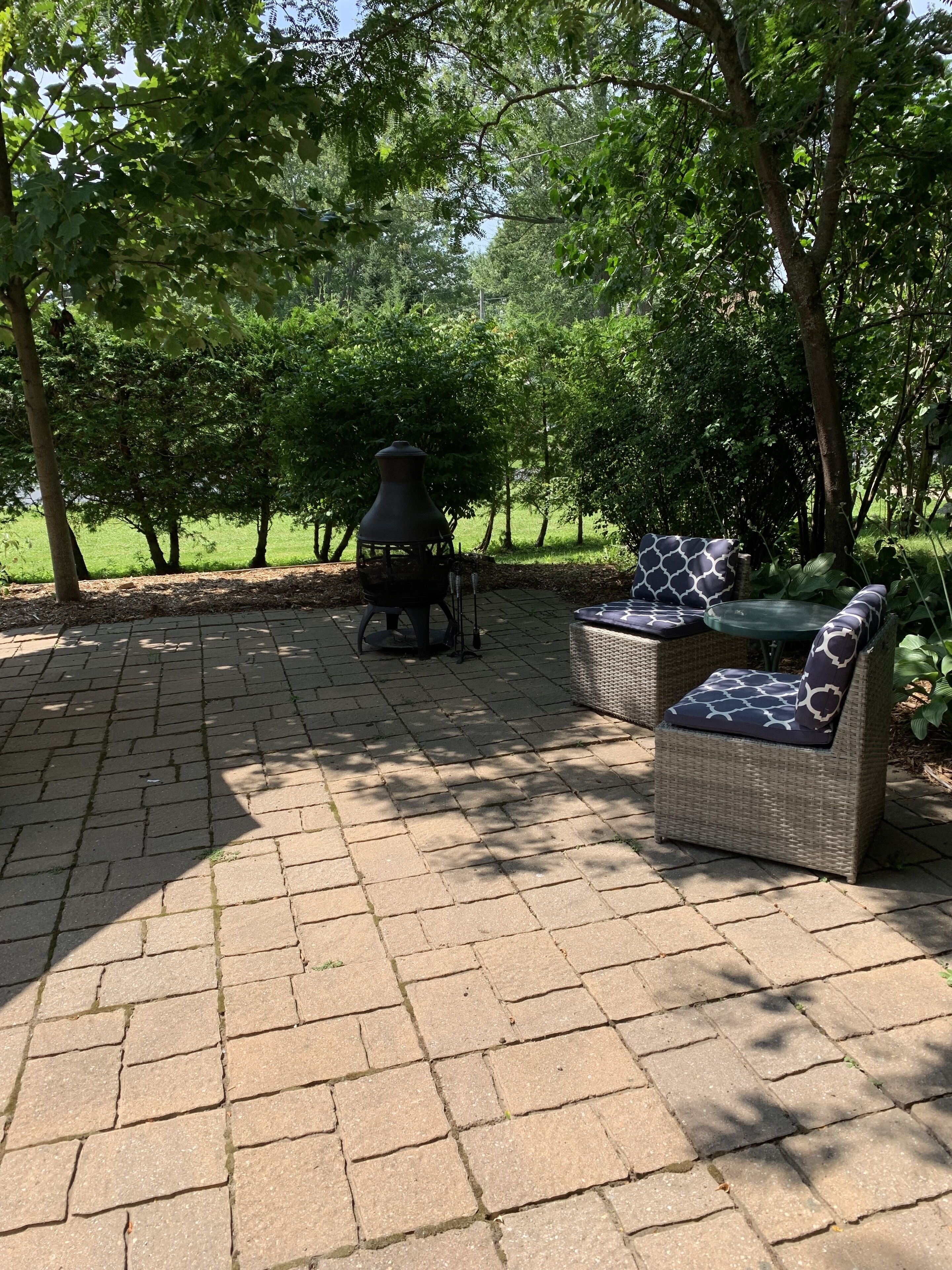 Een terras buiten met een stenen vloer, twee rieten stoelen met opvallende kussens, en een zwarte barbecue.