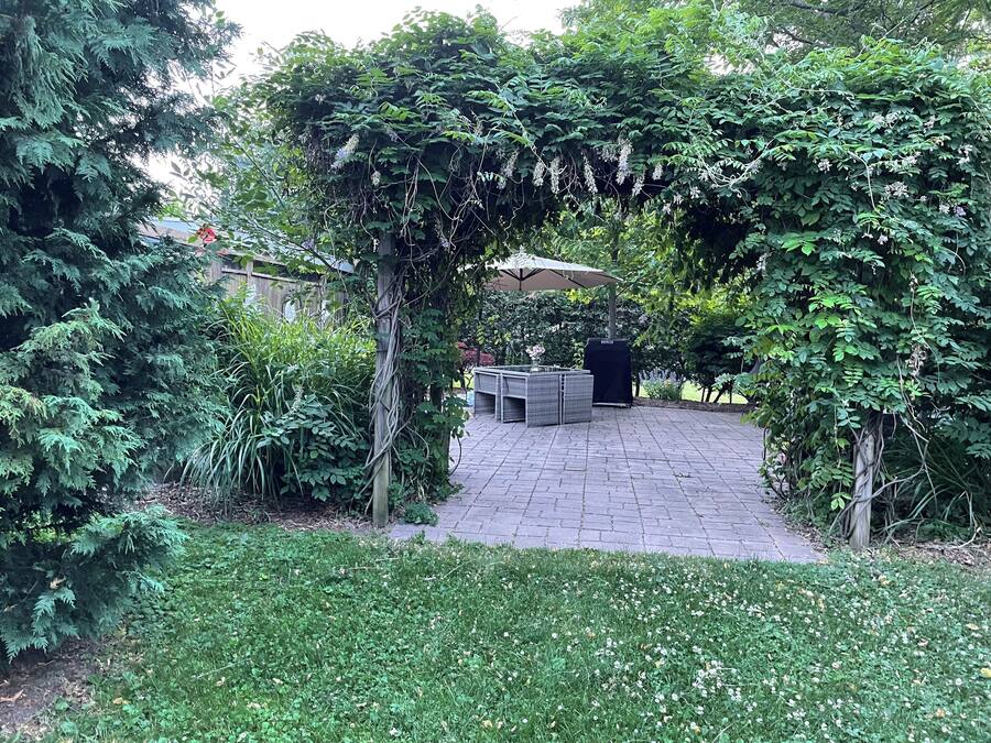 Un patio jardín con piso de ladrillo, rodeado de follaje denso y una pérgola con flores colgantes.