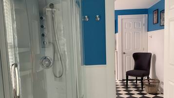 Un baño con cabina de ducha de vidrio, piso a cuadros blanco y negro, una silla oscura y una puerta blanca.