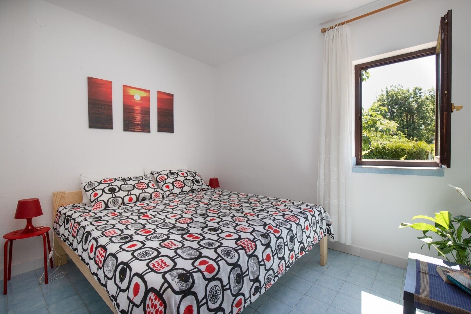 A bedroom with a bed, a nightstand, a window with curtains, and a view of greenery outside.