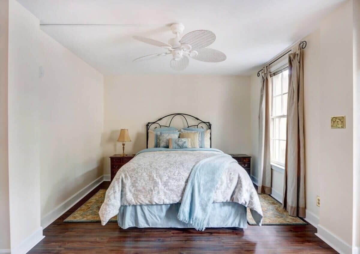 Ein Schlafzimmer mit einem Bett, einem Deckenventilator, einem Nachttisch, einer Lampe und einem Fenster mit Vorhängen.
