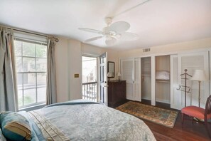 Une chambre à coucher comprenant un lit, un ventilateur de plafond, une fenêtre avec des rideaux, une porte donnant sur l’extérieur, un placard avec des portes coulissantes et une chaise.