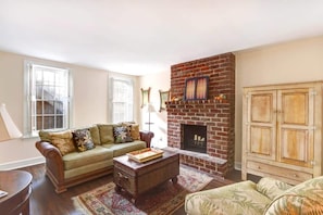 A living room with a brick fireplace, a green sofa, a wooden coffee table, and a patterned rug. - Charming 1800s Garden Apt. | Dog Friendly + Garage (Savannah)