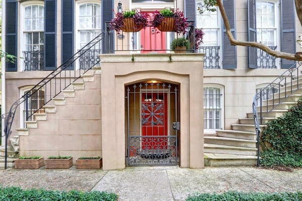 A building with a red door, a staircase, and a balcony with hanging planters. - Charming 1800s Garden Apt. | Dog Friendly + Garage (Savannah)