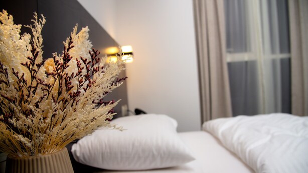 Quarto com cama, almofadas brancas, um vaso com flores secas e luminária fixada na parede.