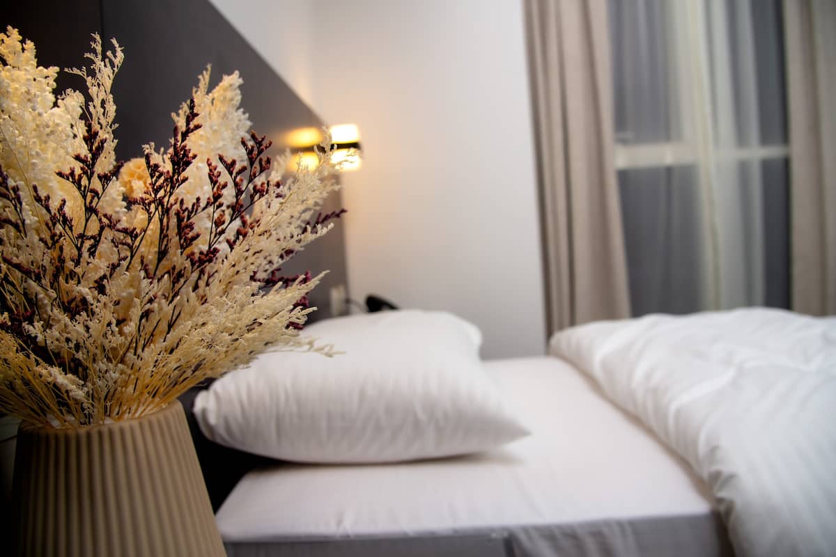 Een slaapkamer met een bed, witte kussens, een vaas met gedroogde bloemen en een wandlamp.