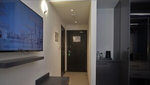 Quarto de hotel com cama, escrivaninha, cadeira, televisão na parede, porta e área de kitchenette.