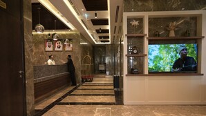 Um saguão de hotel com piso de mármore, um balcão de recepção, uma tela de televisão e um corredor com quadros emoldurados.